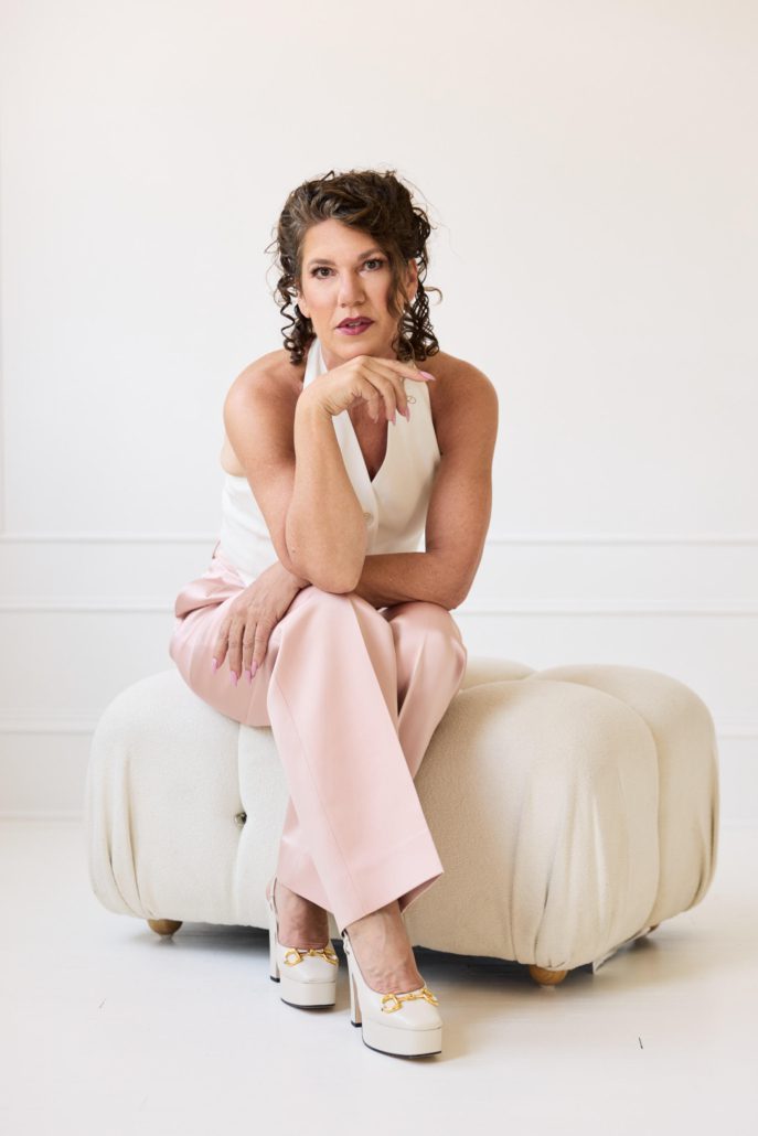 Adele Tosh, founder of Dervilia Art + Design, seated on a sculptural upholstered ottoman in a minimalist studio setting, reflecting her philosophy of meaningful interiors, collected objects, and art-driven design.