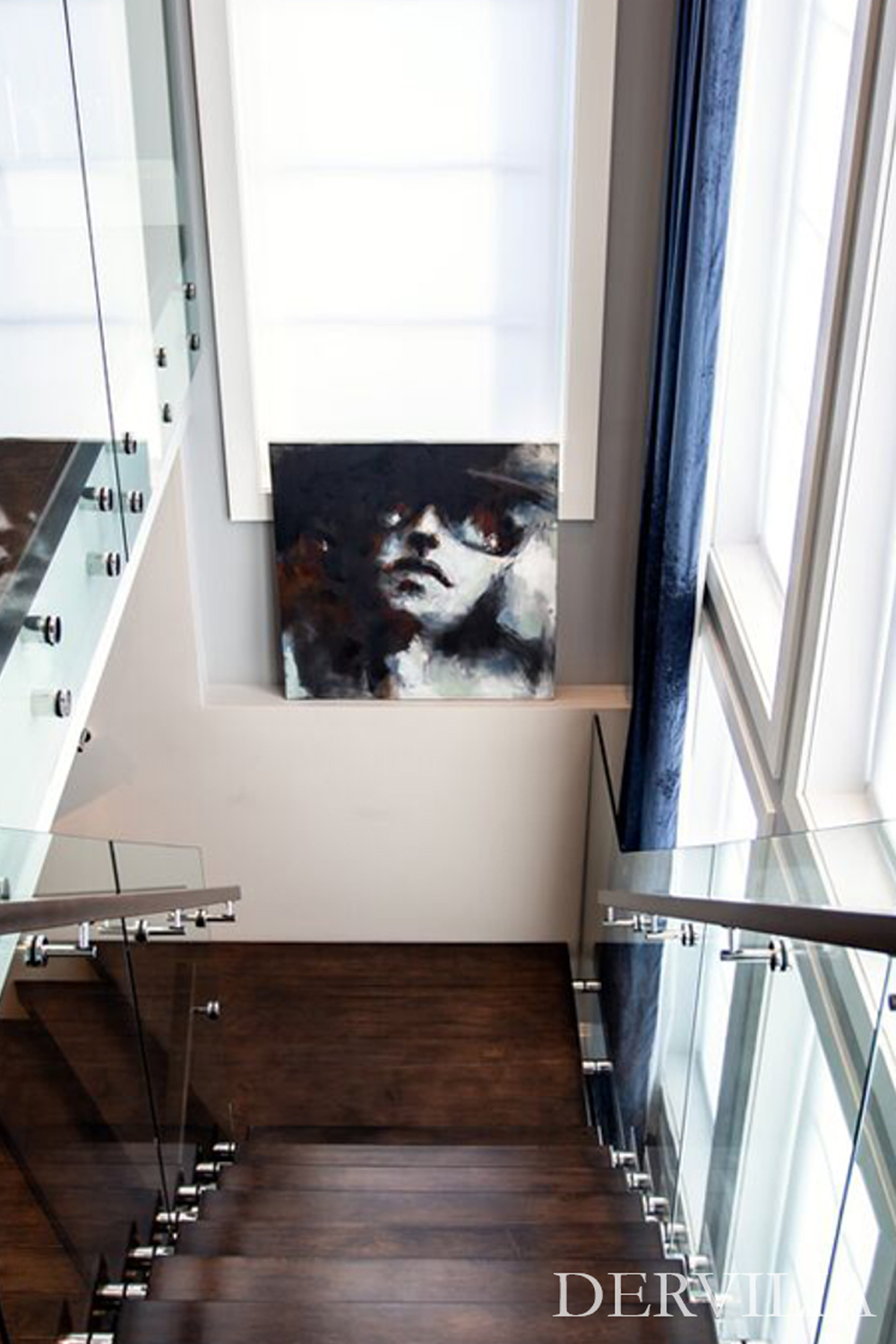 Contemporary staircase with dark wood treads and glass railings, illuminated by tall windows and centered around a dramatic portrait artwork, showcasing Dervilia’s integration of art within architectural design.