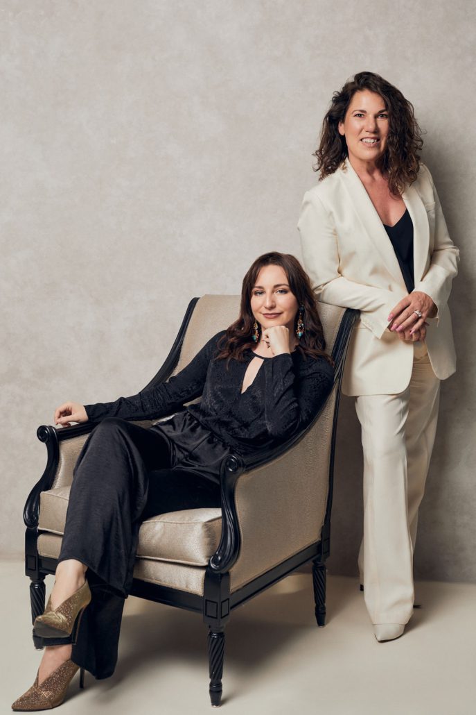 Portrait of two women founders of Dervilia Art + Design, one seated in a classic armchair and the other standing beside her, styled in modern neutral tones that reflect the studio’s contemporary art and interior design philosophy.