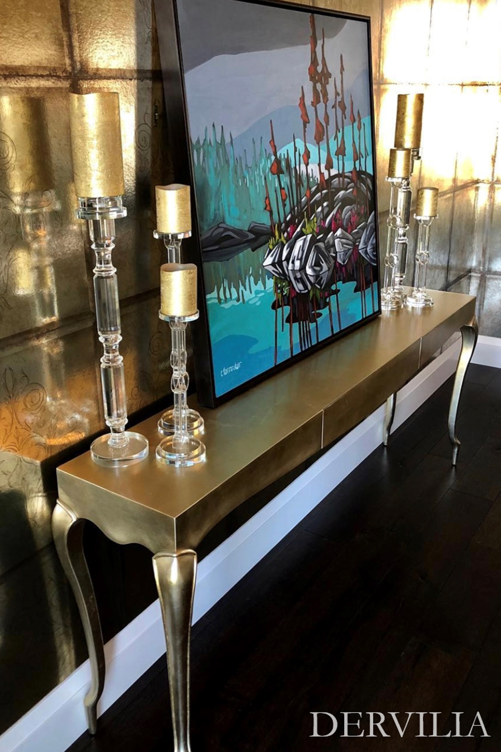 Abstract framed artwork displayed on a metallic console table, styled with tall glass and gold candle holders against a textured wall, highlighting Dervilia’s art-led approach to reflective surfaces and curated interior moments.